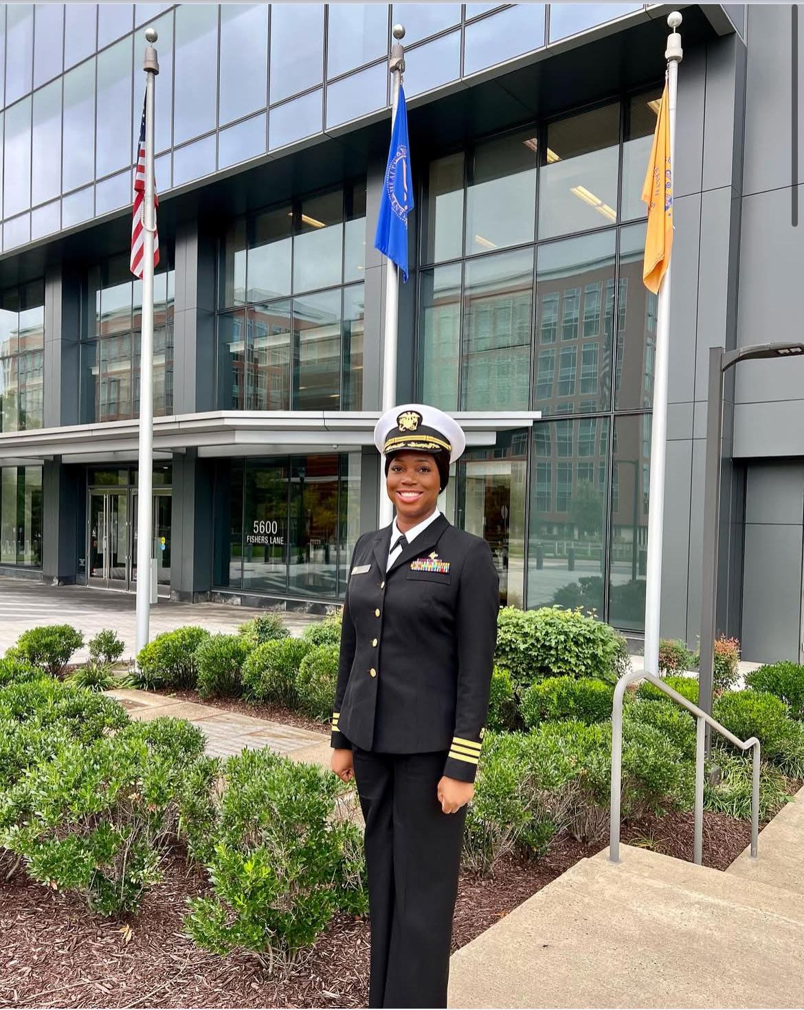 Oye in a dark service uniform standing outside a building with flags.