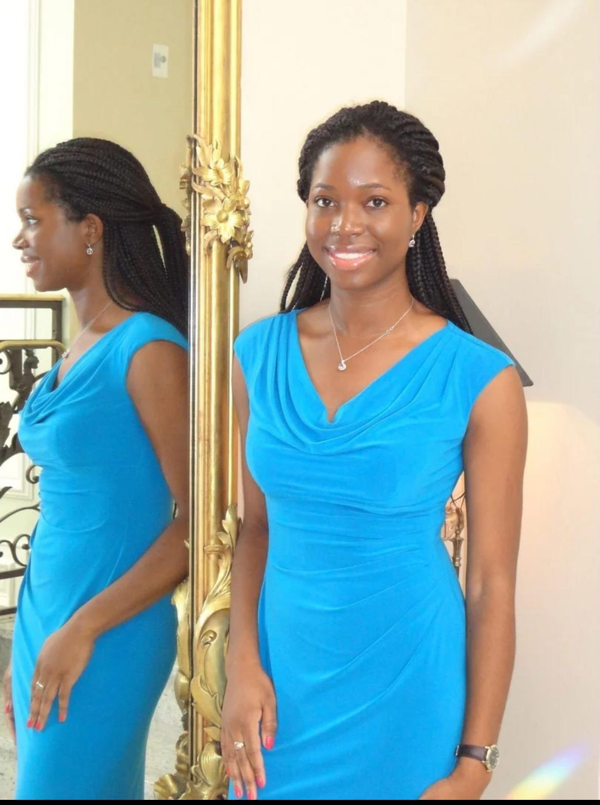 Oye in a blue dress standing beside a tall mirror.
